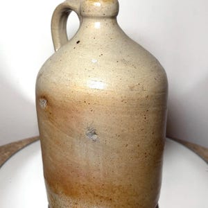 May include: A vintage stoneware jug with a handle. The jug has a rounded body and a narrow neck with a small opening. The exterior is a mottled beige color with brown and tan accents, and a few small imperfections.