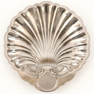 May include: A polished silver-toned metal dish shaped like a large scallop shell. The dish features deep, radiating fluted grooves extending from a smooth, rounded base with two small scroll-like accents near the bottom edge and a scalloped rim.