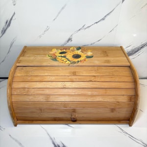May include: A bamboo bread box with a roll-top lid. The box is made of light brown bamboo slats and features a sunflower design on the top. The sunflower design includes yellow, orange, and green colors. A small wooden knob is on the front.