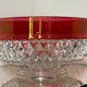 May include: A clear glass bowl with a ruby red rim and a diamond-cut pattern on the lower half. The bowl sits on a clear, circular base, reflecting light. This decorative bowl is ideal for serving or display.