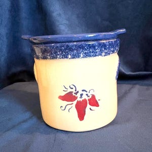 May include: A ceramic crock with a cream-colored body and a blue rim. The crock features a red chili pepper design and a blue handle. The crock is likely used for kitchen storage or decoration.