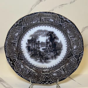 May include: A decorative plate with a black and white landscape scene in the center. The plate has a gray border with ornate detailing and a gold rim. The plate is displayed on a silver metal stand.