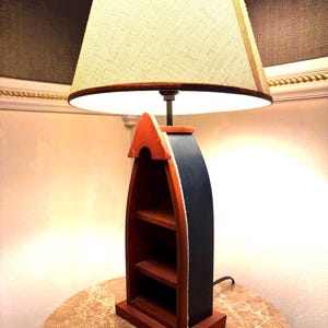 May include: A wooden boat-shaped lamp with a beige lampshade. The lamp base is painted in red, black, and orange, with two shelves inside. The lamp is on a marble-like table.