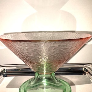 May include: A clear glass bowl with a textured surface and a flared, inverted cone shape. The rim is a reddish-pink color, transitioning to clear in the center. It sits on a green, circular base. The bowl is likely for serving or display.