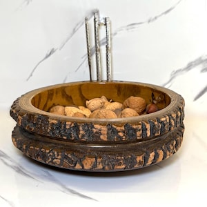 May include: A wooden nut bowl with a rustic, bark-like edge, filled with various nuts. A silver nutcracker stands upright behind the bowl. The bowl is brown and sits on a white surface.
