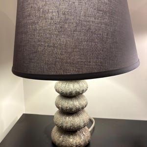 Op de afbeelding: Een decoratieve tafellamp met een gestapelde, getextureerde voet en een grijze stoffen lampenkap. De voet heeft een uniek, gelaagd ontwerp en de lampenkap is een gedempte grijze kleur. De lamp is ontworpen voor gebruik binnenshuis en voegt elegantie toe.