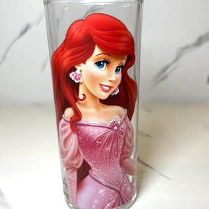 May include: A clear drinking glass decorated with a cartoon character illustration. The character has red hair, blue eyes, and wears a pink dress and earrings. The glass is cylindrical and suitable for beverages.