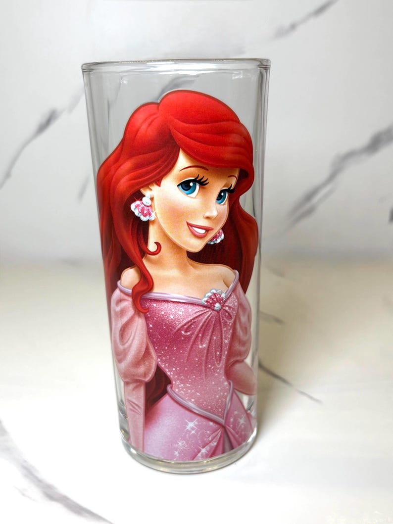 May include: A clear drinking glass decorated with a cartoon character illustration. The character has red hair, blue eyes, and wears a pink dress and earrings. The glass is cylindrical and suitable for beverages.