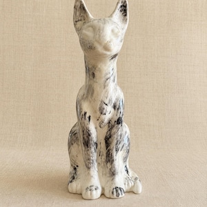 May include: A ceramic cat figurine in a seated position, featuring a glossy white base coat with abstract, hand-painted black and grey brushstroke markings across its entire surface, including pointed ears and a smooth, featureless facial area.