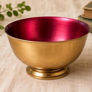 May include: A round bowl with a metallic gold exterior and a vibrant magenta interior. The bowl features a smooth, curved body that tapers down to a circular pedestal base, providing a stable foundation for this decorative metal vessel.