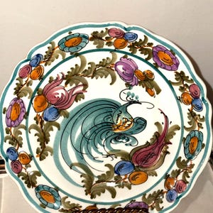 Puede incluir: Un plato de cerámica blanca con borde festoneado y borde turquesa. El plato presenta un diseño central de un pájaro turquesa y motivos florales en naranja, morado y azul. El plato está sostenido por un soporte metálico decorativo.