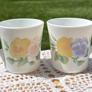 May include: Two white ceramic mugs with floral designs. Each mug features a handle and is decorated with yellow, pink, and purple pansies and green leaves. The mugs are placed on a white, crocheted surface.