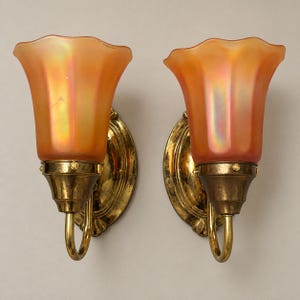 May include: A pair of wall sconces featuring brass backplates and curved arms supporting fluted, iridescent orange glass shades. The metal components exhibit a polished finish with visible patina, while the glass shades have a soft, glowing appearance.