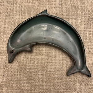 May include: A ceramic dolphin-shaped dish in shades of teal and gray. The dish has a curved design with a detailed dolphin head and tail. The dish is likely used for decorative purposes or as a small serving dish.