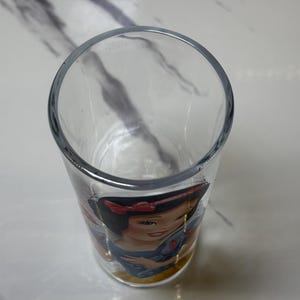 May include: A clear glass tumbler with a Snow White illustration. The glass has a rounded top and a straight, cylindrical body. The Snow White image includes her signature red bow and blue dress. The glass is suitable for drinking.