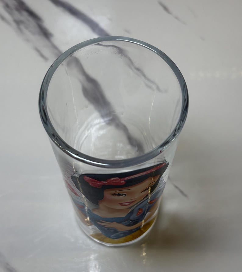 May include: A clear glass tumbler with a Snow White illustration. The glass has a rounded top and a straight, cylindrical body. The Snow White image includes her signature red bow and blue dress. The glass is suitable for drinking.