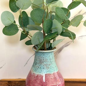 May include: A ceramic vase with a two-tone design, featuring a speckled turquoise top and a reddish-pink base. The vase holds faux eucalyptus branches with round, green leaves. The vase is displayed on a wooden surface.
