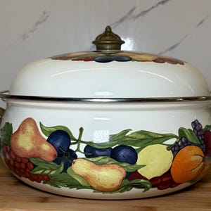 May include: A white enamel cooking pot with a lid, adorned with a colorful fruit design. The pot features gold-colored handles and a silver rim. The fruit illustrations include pears, plums, and oranges. The pot is sitting on a wooden cutting board.
