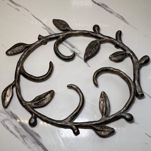 Puede incluir: Una corona decorativa de metal con acabado plateado. El diseño circular presenta hojas estilizadas y ramas curvas. Las hojas tienen una apariencia texturizada. El diseño es simétrico y decorativo.