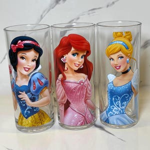 May include: Three clear glass tumblers featuring Disney princesses Snow White, Ariel, and Cinderella. Each tumbler has a different princess illustration in vibrant colours. The glasses are cylindrical and appear to be made of clear glass.