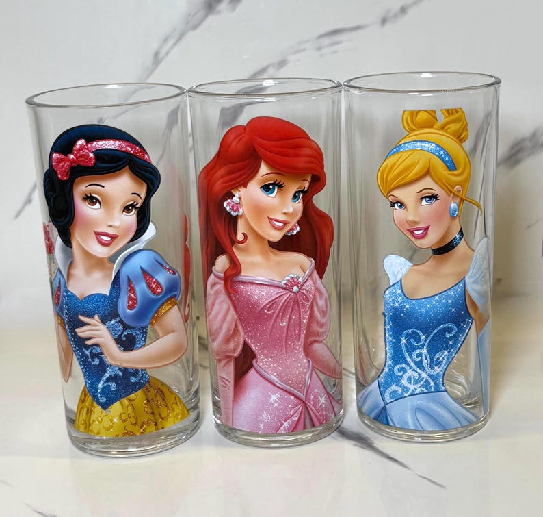 May include: Three clear glass tumblers featuring Disney princesses Snow White, Ariel, and Cinderella. Each tumbler has a different princess illustration in vibrant colours. The glasses are cylindrical and appear to be made of clear glass.