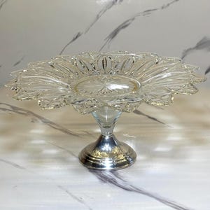 May include: A clear glass cake stand with a decorative, flower-shaped top. The stand has a silver-colored base and stem. The glass top features a detailed, textured design. The stand is designed to display cakes and other desserts.