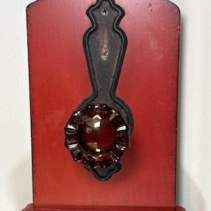 May include: A decorative wall-mounted piece with a red wooden backing and a dark metal accent. It features a brown, flower-shaped glass knob. The piece is designed for wall mounting and has a shelf-like base.