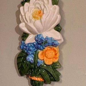 Puede incluir: Adorno floral de cerámica para colgar en la pared. Presenta una gran flor blanca con centro amarillo, rodeada de flores azules y naranjas y hojas verdes. Las flores están dispuestas verticalmente, con un acento naranja arriba y abajo.