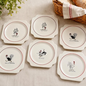 May include: A set of six square white ceramic plates with rounded corners, each featuring a thin red circular border and a unique black and white illustration of a rooster with a small red accent, accompanied by descriptive text below each image.