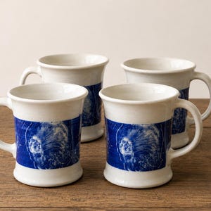 May include: A set of four off-white ceramic mugs, each featuring a wide, horizontal band of deep blue glaze around the center. The blue band is decorated with a white, intricate, swirling graphic pattern that resembles a celestial or nebula design.