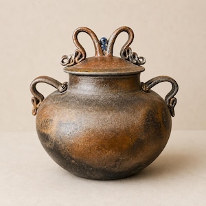 May include: A round, dark brown and black ceramic jar with a textured, mottled finish. It features two curved side handles and a matching lid topped with an ornate, looped handle design that includes a small, round, blue stone accent at the center.