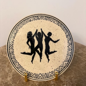 Puede incluir: Un plato redondo de cerámica con un fondo beige moteado. El plato presenta un borde negro con un diseño geométrico y una silueta central de tres figuras bailando. El plato se exhibe sobre un soporte dorado.