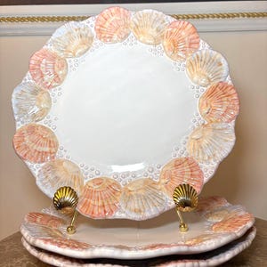 May include: White ceramic plates with a seashell design around the rim. The seashells are in shades of peach, pink, and white. The plates are stacked on a brown surface.