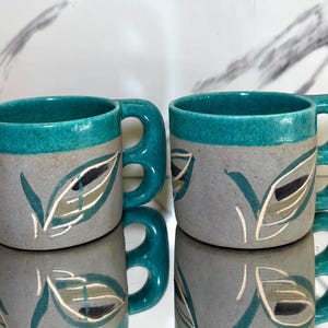 May include: Two ceramic mugs with teal rims and handles. The mugs are gray with a leaf design in teal, black, and white. The mugs are reflected in the surface below.