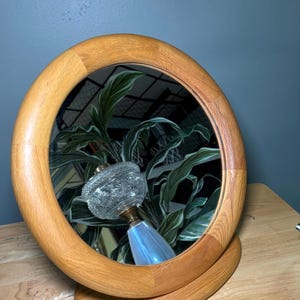 May include: A round wooden vanity mirror with a light brown finish. The mirror reflects a clear glass perfume bottle with a decorative stopper. The mirror is on a wooden base.