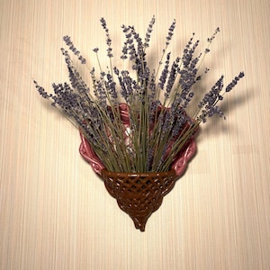 May include: A wall-mounted decorative piece featuring a bouquet of dried lavender. The lavender is arranged in a brown, woven basket-like holder, set against a pink, scalloped backing. The piece is mounted on a textured, light-colored wall.