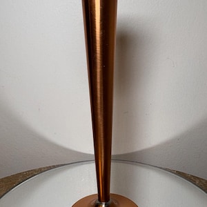May include: A copper-colored bud vase with a flared top and a round base. The vase has a long, slender body and is made of a shiny metal. The vase is sitting on a reflective surface.