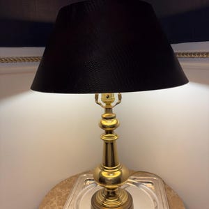 May include: A brass table lamp with a black lampshade. The lamp has a detailed, ornate base and a textured lampshade. The lamp is sitting on a marble-topped table with a silver tray.