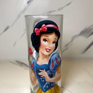 May include: A clear glass with a vibrant depiction of Snow White. The design showcases her black hair, red bow, blue bodice, and yellow skirt. The cylindrical glass is suitable for drinks and beverages.