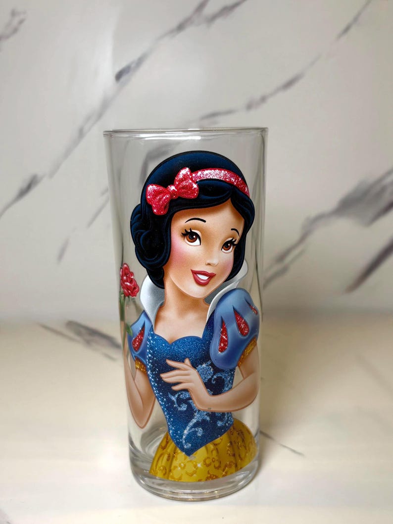 May include: A clear glass with a vibrant depiction of Snow White. The design showcases her black hair, red bow, blue bodice, and yellow skirt. The cylindrical glass is suitable for drinks and beverages.