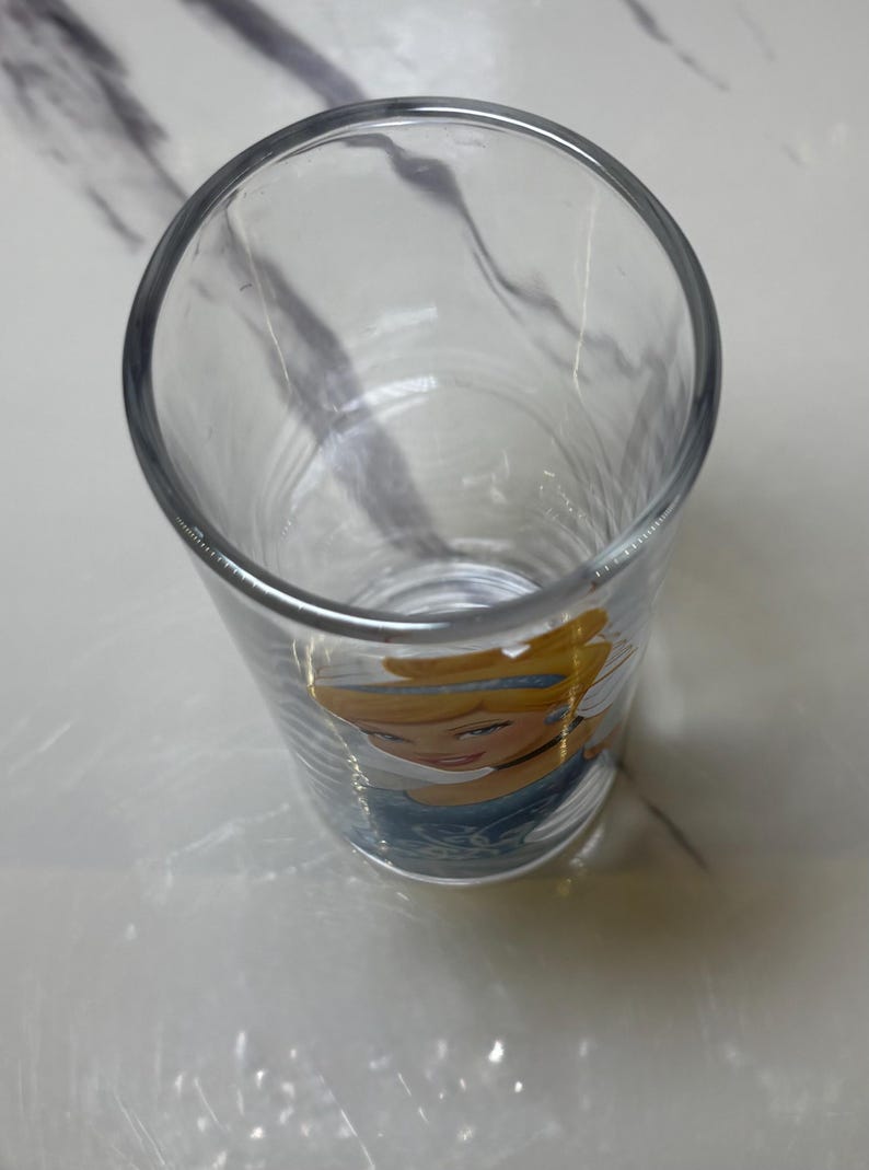 May include: A clear glass tumbler featuring a cartoon image of Cinderella. The glass has a cylindrical shape and a thick rim. The image of Cinderella is in the lower portion of the glass, with her face and upper body visible.