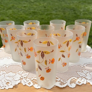 May include: A set of ten frosted glass tumblers. Each glass features a floral design with orange flowers and brown and yellow leaves. The glasses are arranged on a white lace tablecloth, with a green grassy background.