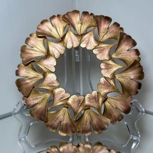 Puede incluir: Una corona decorativa con un diseño circular de hojas de ginkgo estilizadas. Las hojas tienen un acabado metálico con tonos dorados y bronce. La corona se exhibe sobre un soporte transparente.