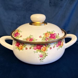 May include: White enamelware pot with lid, featuring a floral rose pattern in shades of pink, yellow, and green. The pot has two handles and a gold-toned rim and lid accents. A classic kitchenware item.