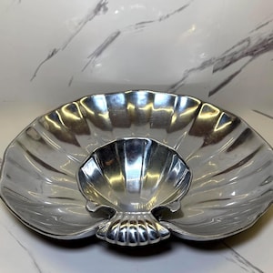 May include: A silver-colored, shell-shaped serving dish with a smaller, matching dish nested inside. The larger dish has a scalloped edge and a textured surface. The smaller dish is also shell-shaped and sits on a decorative base.