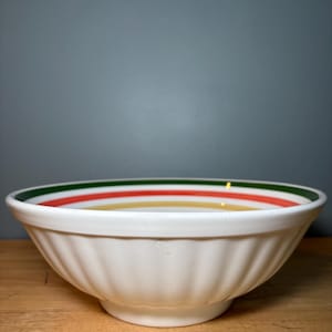 May include: White ceramic bowl with a fluted exterior and a colorful striped rim. The rim features green, red, and yellow bands. The bowl sits on a wooden surface against a gray background. A classic kitchenware item.