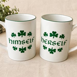 May include: Two white ceramic mugs with a dark green rim. Each mug features a circular pattern of green shamrocks surrounding the word "himself" on one mug and "herself" on the other, both written in a stylized, bold, dark green font.