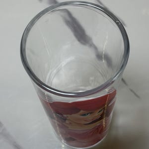 May include: Clear drinking glass with a colourful image of Ariel from The Little Mermaid. The glass has a wide opening and a slightly tapered design. The image of Ariel is in shades of red, pink, and green.