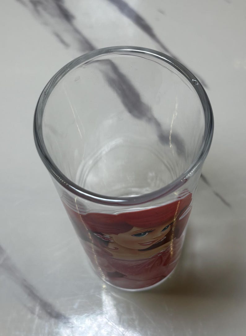 May include: Clear drinking glass with a colourful image of Ariel from The Little Mermaid. The glass has a wide opening and a slightly tapered design. The image of Ariel is in shades of red, pink, and green.