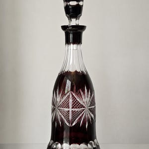 May include: A tall, deep red and clear cut glass decanter featuring a matching stopper. The body displays intricate starburst and diamond cross-hatch patterns, with a series of clear circular cutouts along the base and a fluted neck design.